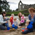 Young children playing on the beach area next to the River Wharfe at Bolton Abbey. The priory ruins provide a stunning backdrop.