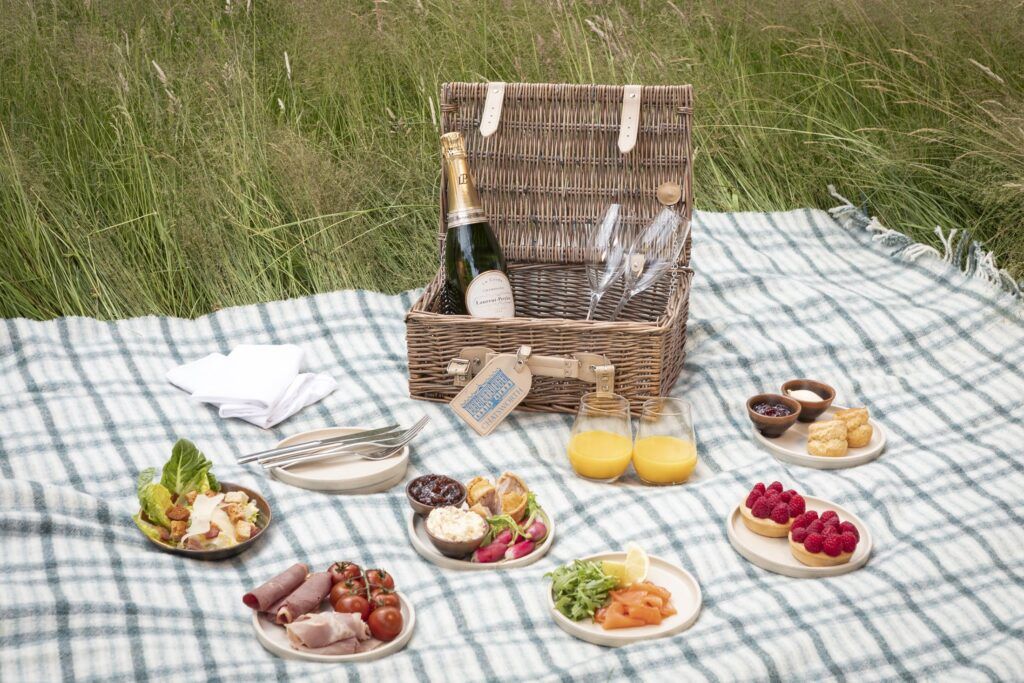 A picnic basket placed on a rug out in the Bolton Abbey countryside with a bottle, glasses and a selection of food